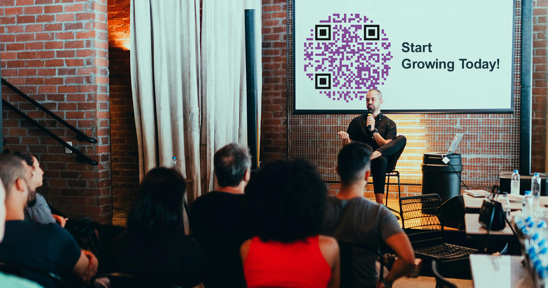 Conference with speaker at the front. Behind the speaker is a large screen with a QR Code next to the text : Start Growing Today!